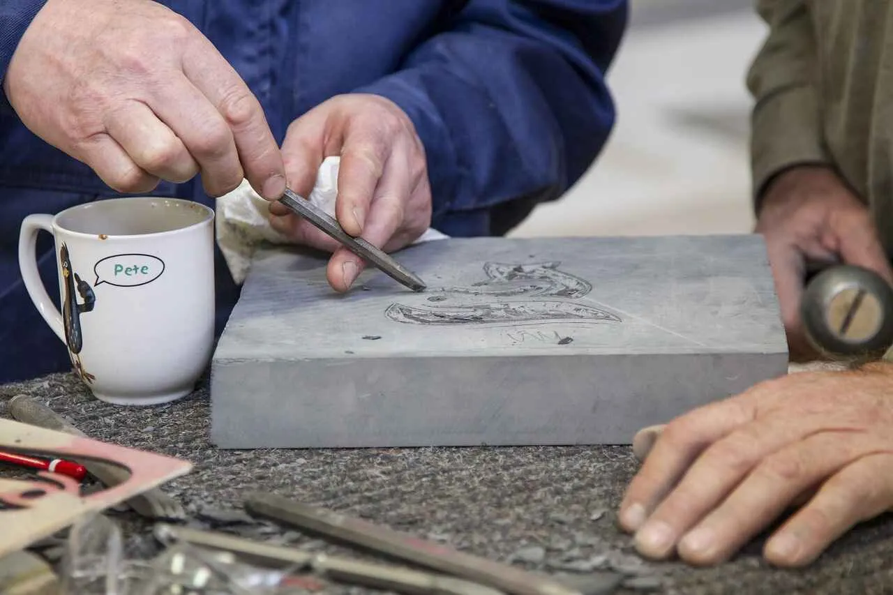 A woman is working on a piece of slate with a chisel.