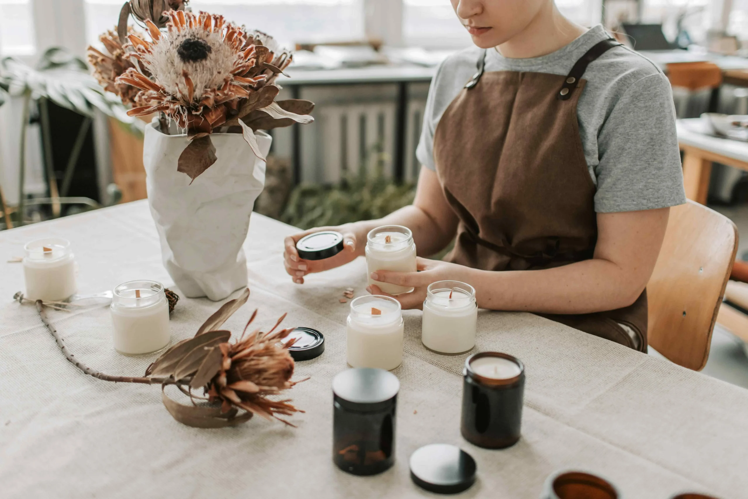 A woman in an apron is carefully arranging candles on a table, with dried flowers in a vase nearby.