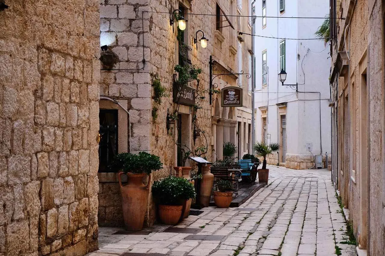 A narrow cobblestone street in a European town, with potted plants and a restaurant with outdoor seating.