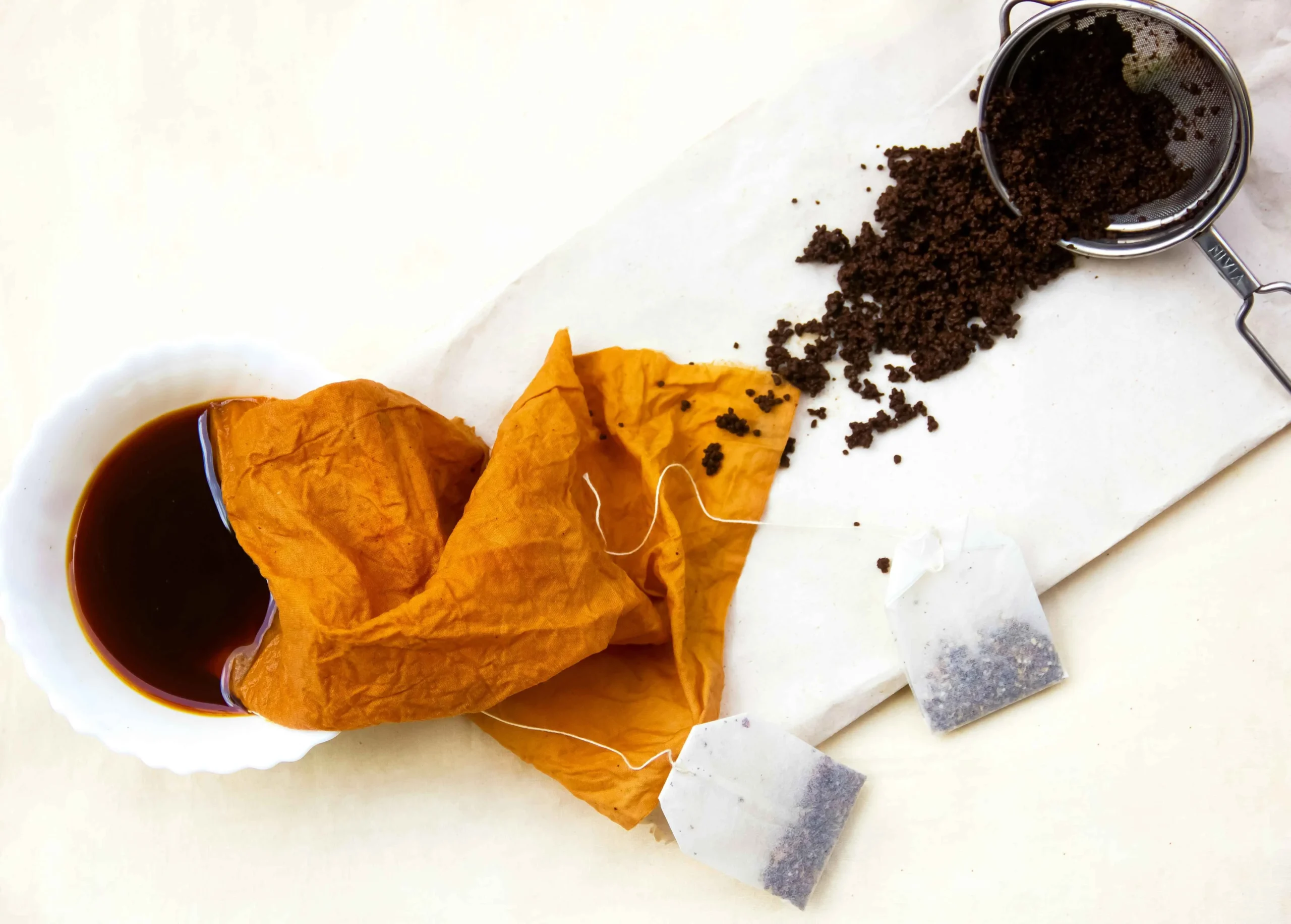 A close-up of natural dyeing a white bowl of dark liquid, golden-brown cloth, tea bag, loose tea leaves, and a white surface.